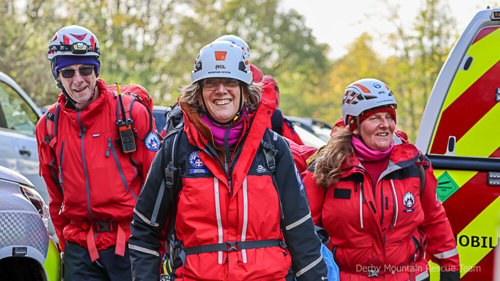 Three smiling mountain rescue volunteers in helmets walking past a team vehicle